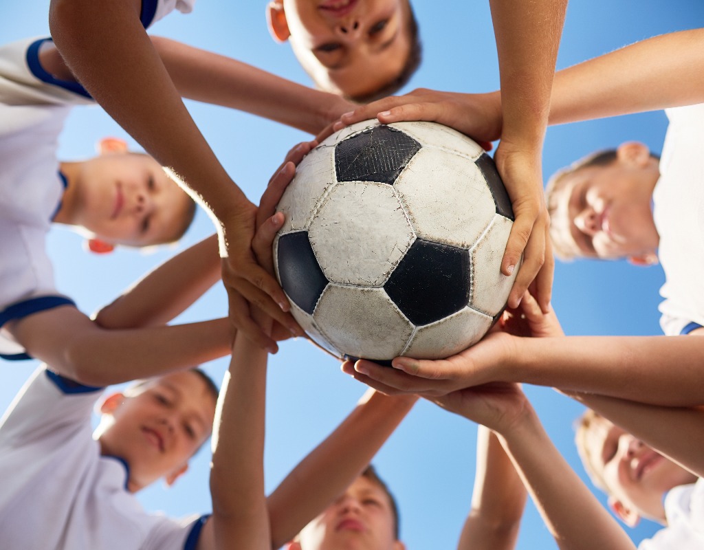 Low angle view of boys in junior football team standing in circle holding ball together against  blue sky, focus on ball
