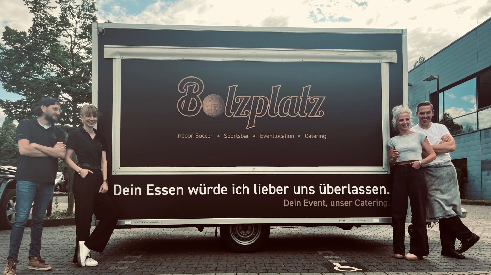 Zwei Paare umarmen sich lächelnd vor einem schwarzen Catering-Fahrzeug.