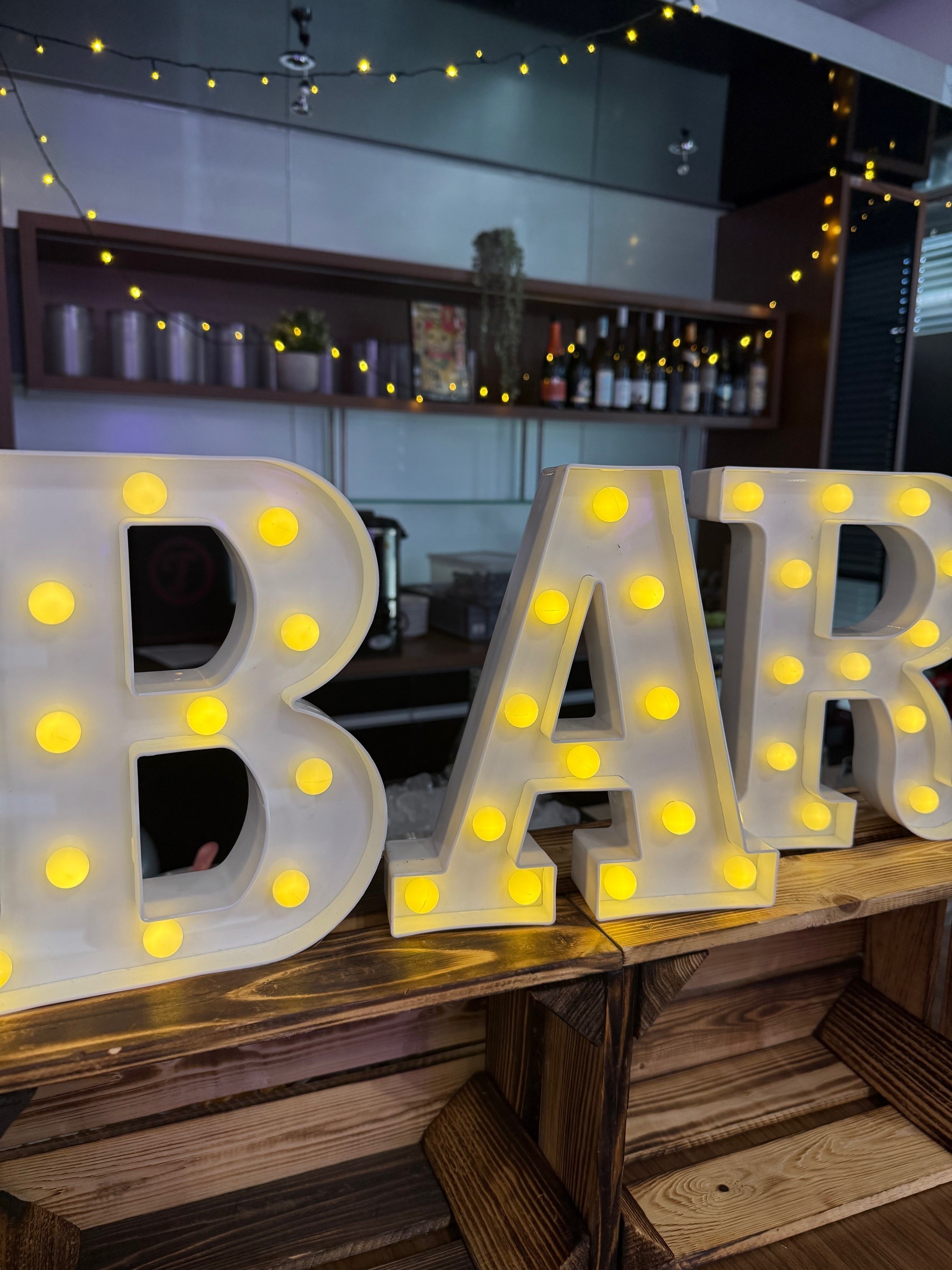 Bar-Schild mit gelben Leuchten; im Hintergrund eine Theke mit Lichtern und Flaschen.