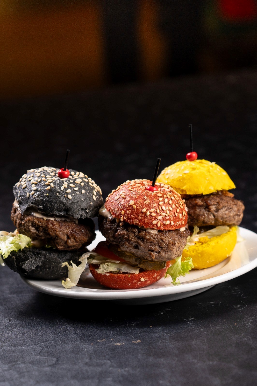 Drei Mini-Burger mit farbigen Brötchen (schwarz, rot, gelb) auf einem Teller, mit Salat verziert.