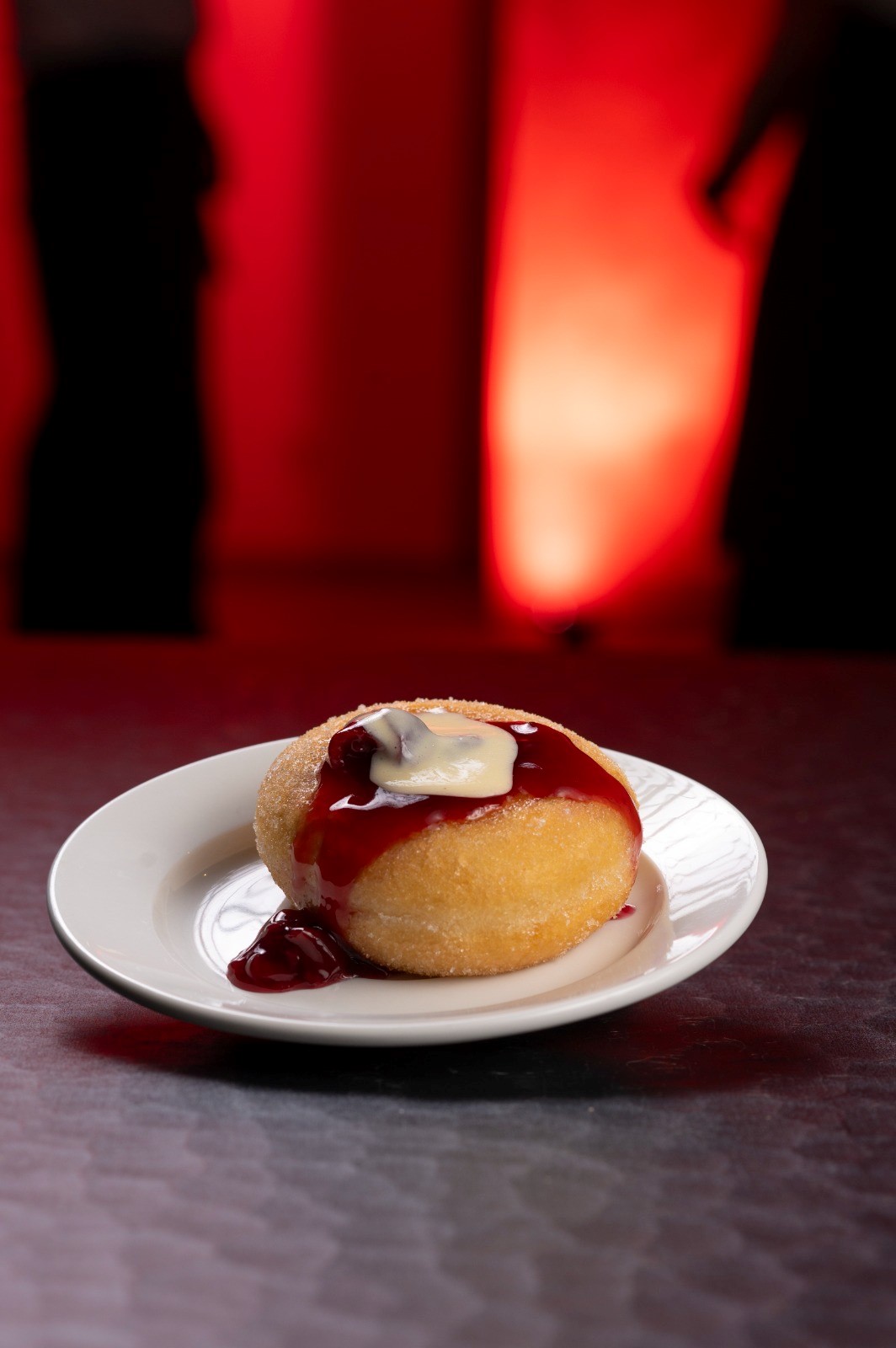 Ein Berliner mit roter Sauce und Sahne auf einem Teller vor rotem Hintergrund.