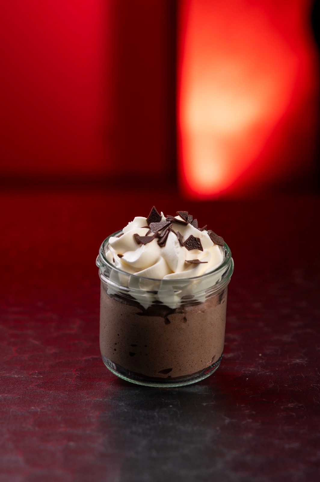 Schokoladenmousse in Glas, garniert mit Sahne und Schokoladenstückchen, roter Hintergrund.