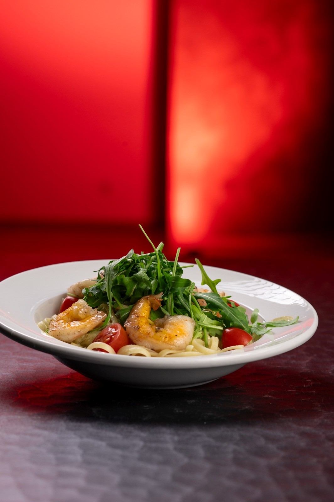 Ein Teller Pasta mit Garnelen, Rucola und Kirschtomaten vor einem roten Hintergrund.
