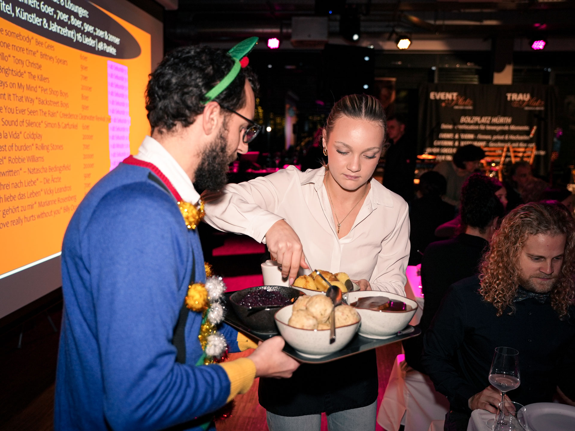 Eine Frau mit blondem Zopf legt konzentriert Essen auf einen Tisch bei warmem Licht