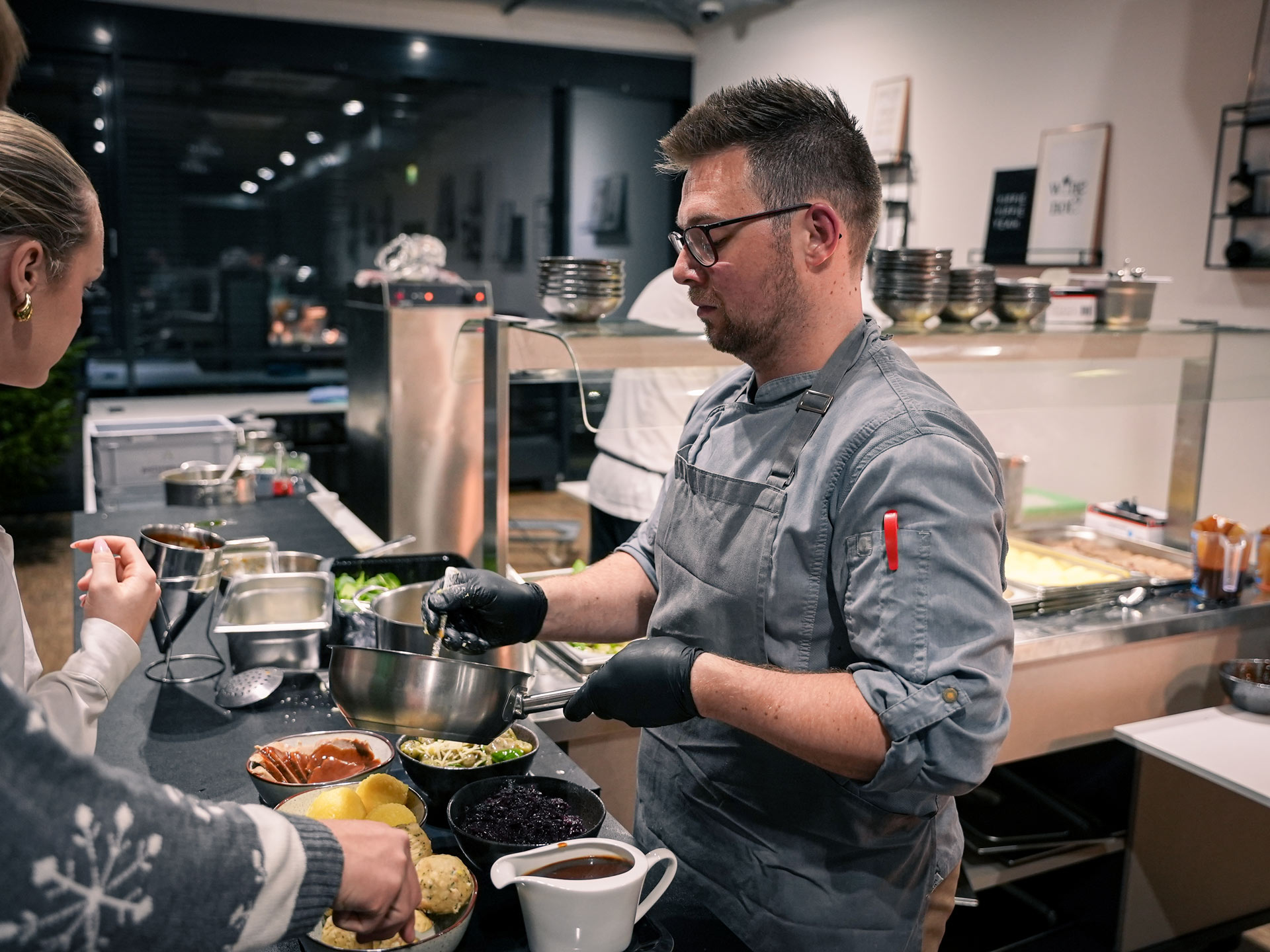 Ein Mann mit Brille und grauer Kochjacke bereitet konzentriert eine Mahlzeit zu