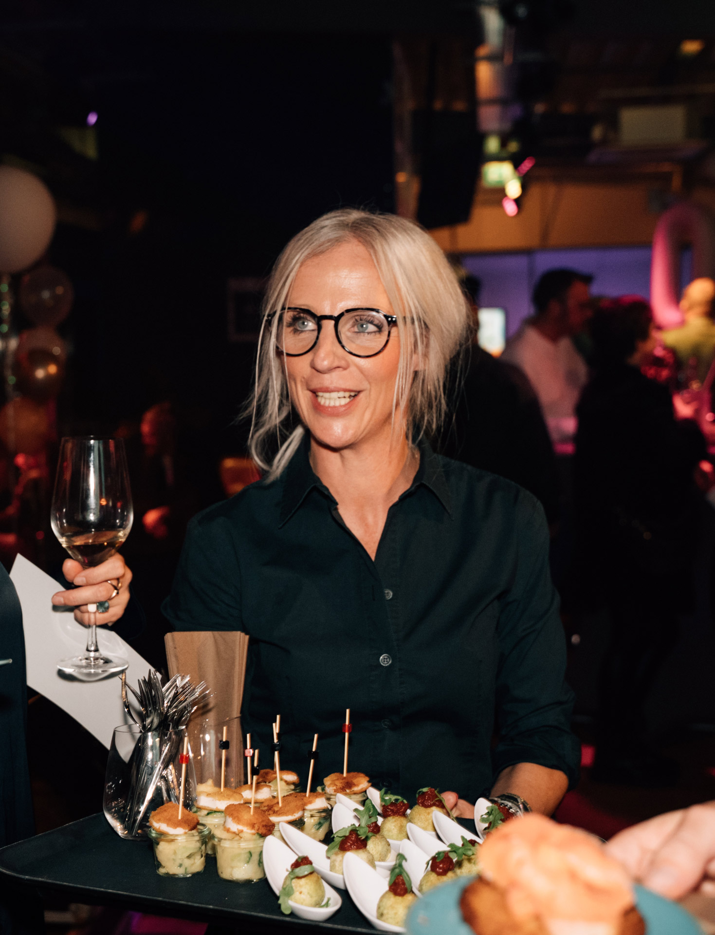 Eine lächelnde Frau mit blonden Haaren und Brille hält ein Glas Wein und einen Teller Snacks
