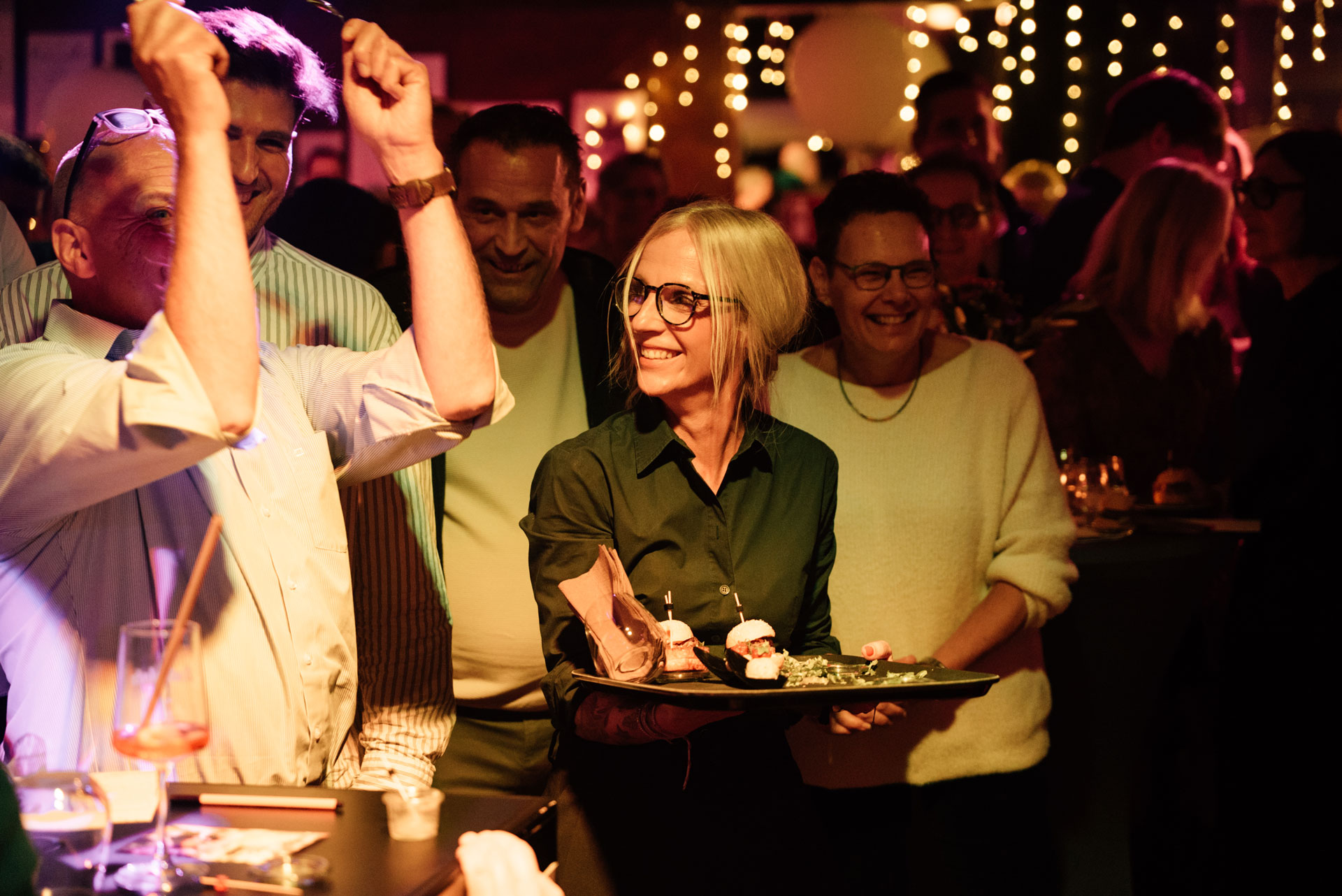 Eine blonde Frau mit Brille lächelt inmitten einer fröhlichen Feiergesellschaft