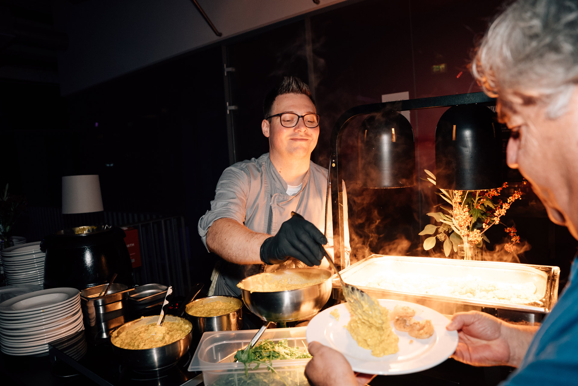 Ein lächelnder Koch mit Brille serviert einem Gast gelbe Speisen am Buffet